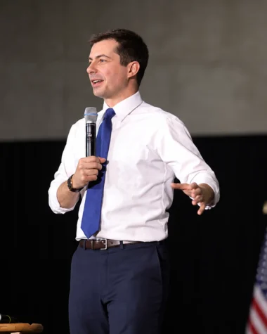 AI-assisted recreation of Pete Buttigieg speaking into a microphone in a white shirt and blue tie, with flags behind him.