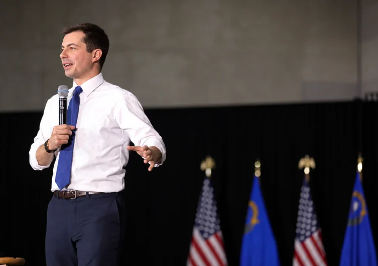 AI-assisted recreation of Pete Buttigieg speaking into a microphone in a white shirt and blue tie, with flags behind him.